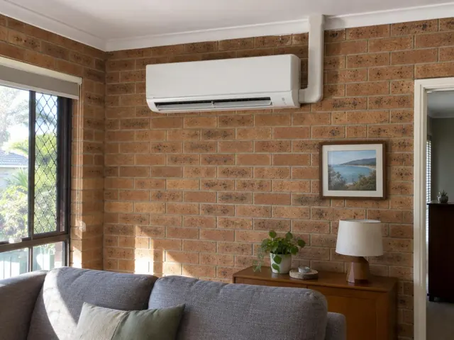 Wall-mounted split system on a brick living room wall with window and furniture in Worongary